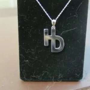 Polished silver pendant featuring the letters "H" and "B" merged into a single monogram design on a black display stand.