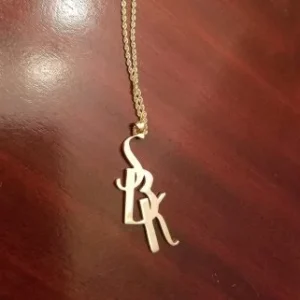 A gold vertical pendant featuring the initials "SBK" in a stylized, interlocking script font, hanging from a gold rope chain against a dark wooden surface.