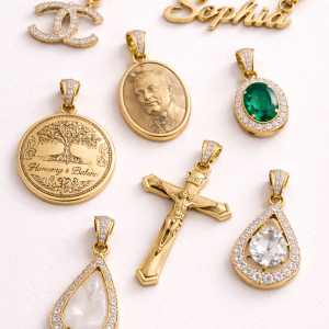 An assortment of eight high-end gold pendants arranged on a white textured background. The collection features a variety of designs including an iced-out interlocking logo, a "Sophia" nameplate, a custom photo-engraved oval medallion, an oval emerald stone with a halo, a "Harmony & Balance" tree of life coin, a detailed gold crucifix, and two teardrop-shaped pendants—one with a clear faceted stone and another with a smooth white inlay, both surrounded by shimmering pavé borders. All pendants include decorative iced-out bails.